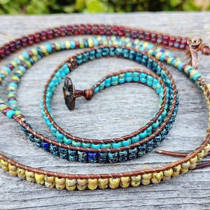 Op de afbeelding: Een wikkelarmband met meerdere strengen, bestaande uit bruin leren koord en diverse kleine glaskralen in tinten turquoise, geel, donkerrood en donkerblauw. De armband heeft een donker metalen bloemvormige knoopsluiting en een klein blaadje.