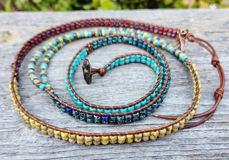Op de afbeelding: Een wikkelarmband met meerdere strengen, bestaande uit bruin leren koord en diverse kleine glaskralen in tinten turquoise, geel, donkerrood en donkerblauw. De armband heeft een donker metalen bloemvormige knoopsluiting en een klein blaadje.