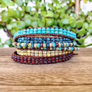 Op de afbeelding: Een wikkelarmband met meerdere strengen, bestaande uit rijen kleine, ronde, gespikkelde kralen in tinten turkoois, donkerblauw, geel en dieprood. De kralen zijn bevestigd met bruin leren koord, wat zorgt voor een gelaagde uitstraling met textuur.