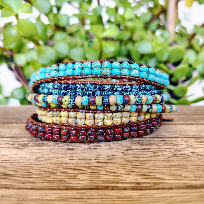 Op de afbeelding: Een wikkelarmband met meerdere strengen, bestaande uit rijen kleine, ronde, gespikkelde kralen in tinten turkoois, donkerblauw, geel en dieprood. De kralen zijn bevestigd met bruin leren koord, wat zorgt voor een gelaagde uitstraling met textuur.