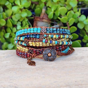 Op de afbeelding: Een wikkelarmband met meerdere strengen, bestaande uit rijen kleine, kleurrijke glaskralen in tinten turquoise, blauw, geel en rood, geregen aan een bruin leren koord. De armband bevat koperkleurige metalen bedels in de vorm van bloemen en een blad.