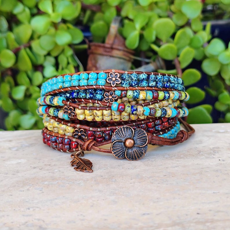 Op de afbeelding: Een wikkelarmband met meerdere strengen, bestaande uit rijen kleine, kleurrijke glaskralen in tinten turquoise, blauw, geel en rood, geregen aan een bruin leren koord. De armband bevat koperkleurige metalen bedels in de vorm van bloemen en een blad.