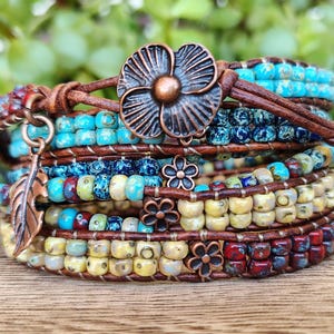 Op de afbeelding: Een meerstrengs wikkelarmband met bruin leren koord, turquoise, blauwe, gele en rode glaskralen en koperkleurige metalen bedels, waaronder een grote bloemvormige knoopsluiting, een bladvormige hanger en drie kleine bloemaccenten.