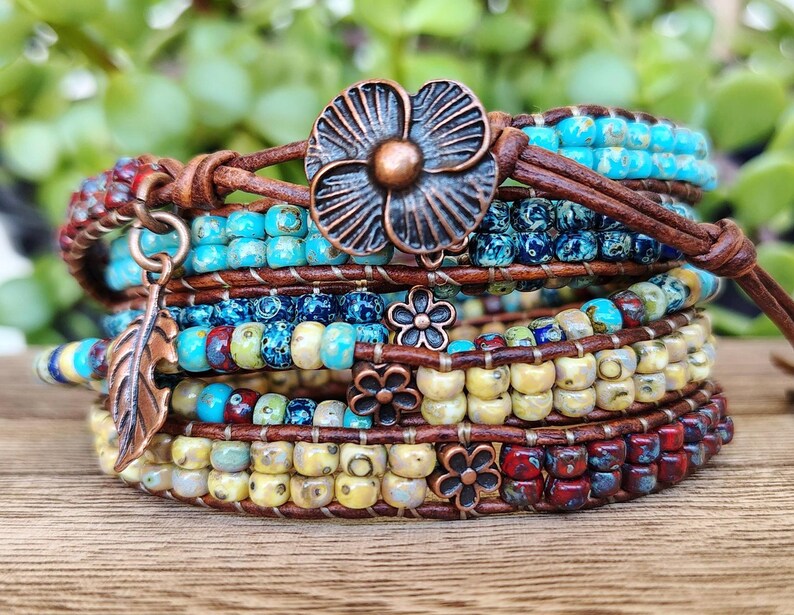 Op de afbeelding: Een meerstrengs wikkelarmband met bruin leren koord, turquoise, blauwe, gele en rode glaskralen en koperkleurige metalen bedels, waaronder een grote bloemvormige knoopsluiting, een bladvormige hanger en drie kleine bloemaccenten.