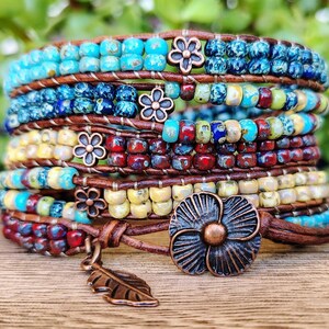 Op de afbeelding: Een wikkelarmband met meerdere strengen, voorzien van rijen kleurrijke kralen in tinten turquoise, blauw, rood en geel, geweven op bruine leren koorden. De armband bevat kleine metalen bloemaccenten en een grote bloemvormige knoopsluiting.