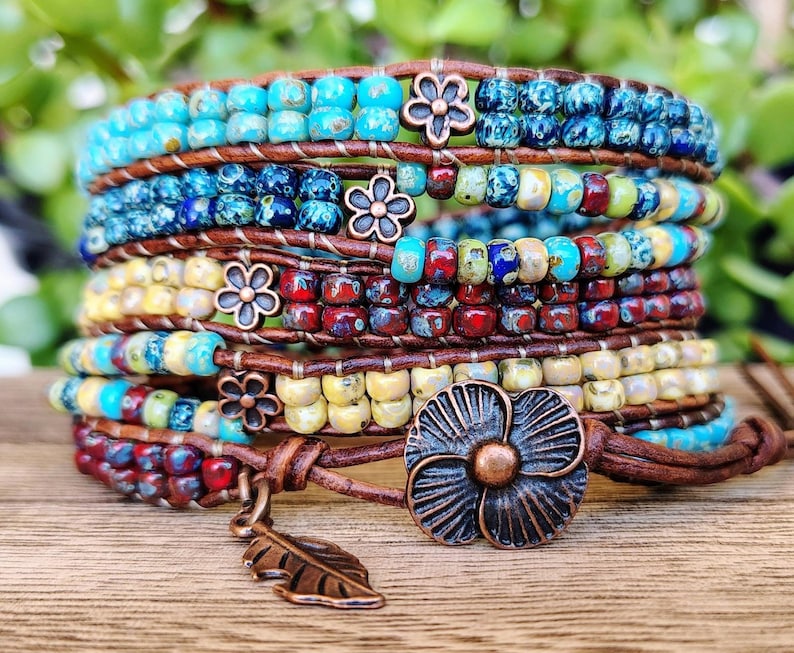Op de afbeelding: Een wikkelarmband met meerdere strengen, voorzien van rijen kleurrijke kralen in tinten turquoise, blauw, rood en geel, geweven op bruine leren koorden. De armband bevat kleine metalen bloemaccenten en een grote bloemvormige knoopsluiting.