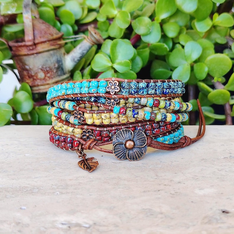 Op de afbeelding: Een wikkelarmband met meerdere strengen, bestaande uit rijen kleine, kleurrijke glaskralen in tinten turquoise, blauw, geel en rood. De armband is versierd met kleine koperkleurige metalen bloembedels en een grote, gedetailleerde bloemvormige knoopsluiting.