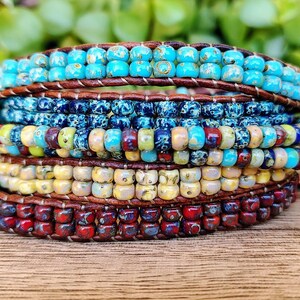 Op de afbeelding: Een wikkelarmband met meerdere strengen, voorzien van rijen kleine, ronde, gevarieerde glaskralen in tinten turquoise, blauw, geel en rood, vastgezet tussen twee donkerbruine leren koorden met zichtbare stiksels over het hele ontwerp.