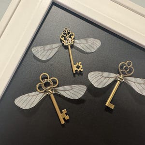 May include: Three antique-style keys with delicate, translucent wings attached, arranged on a dark surface. The keys have ornate designs and a brassy finish, creating a whimsical, decorative effect. The keys are framed in a white picture frame.