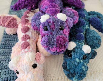 May include: Three crocheted dragon plushies in different colors: pink, purple, and blue. Each dragon has white eyes and a white nose.