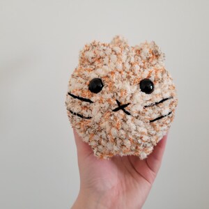 May include: A small, plush cat toy with a white and orange fur pattern. The cat has black eyes and a black stitched nose and mouth.
