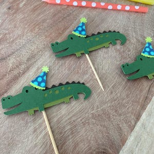 May include: Green alligator cupcake toppers with blue polka dot party hats. The toppers are on wooden sticks, arranged on a wooden surface. Polka dot candles in blue, yellow and orange are in the background.