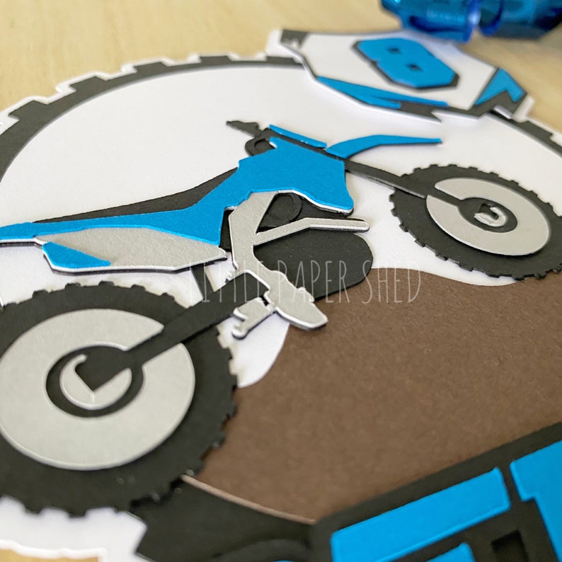 May include: A blue and silver motocross bike with black tires, on a brown background. The bike is partially cut out, with the front wheel and handlebars missing.