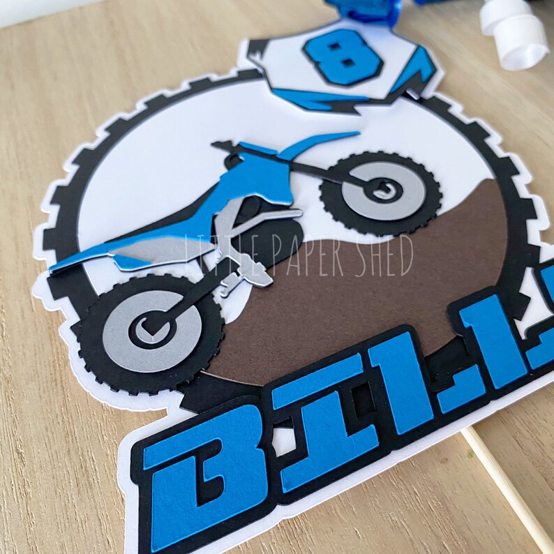May include: A blue and black motocross cake topper with the name "BILL" in blue letters. The motocross bike is jumping over a brown dirt mound. The topper is on a wooden surface.