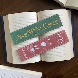 May include: Two rectangular bookmarks are displayed on an open book. One is green with gold text reading "SHOP AROUND THE CORNER." The other is red with a fox illustration and the text "FOX & SONS BOOKS."