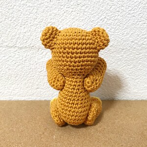 Puede incluir: Un oso de peluche tejido a crochet en posici&oacute;n sentada, de espaldas a la c&aacute;mara. El oso es de color marr&oacute;n claro y tiene un dise&ntilde;o sencillo.