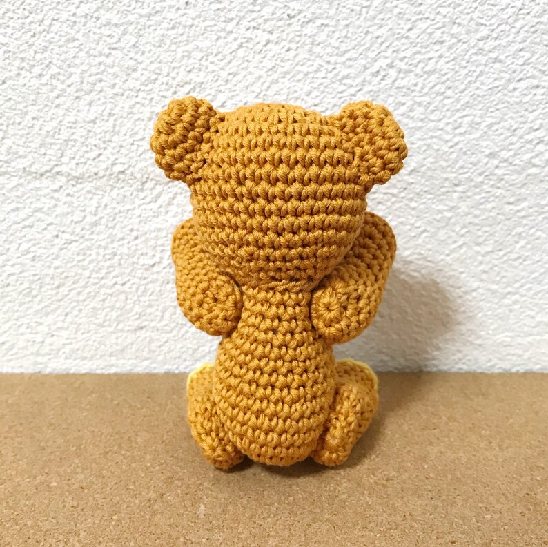 Puede incluir: Un oso de peluche tejido a crochet en posici&oacute;n sentada, de espaldas a la c&aacute;mara. El oso es de color marr&oacute;n claro y tiene un dise&ntilde;o sencillo.