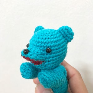 Puede incluir: Un oso de peluche de color turquesa azul tejido a crochet con ojos negros y una boca roja.