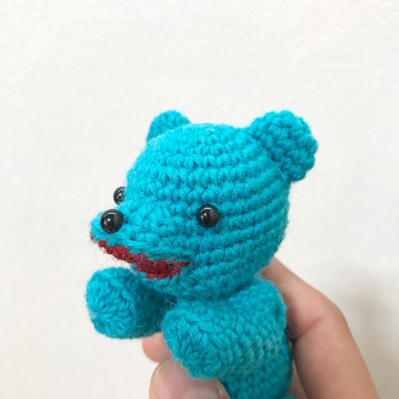 Puede incluir: Un oso de peluche de color turquesa azul tejido a crochet con ojos negros y una boca roja.