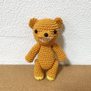 Puede incluir: Un oso de peluche de crochet con cuerpo marr&oacute;n, ojos negros y una sonrisa cosida blanca. El oso est&aacute; de pie sobre una superficie marr&oacute;n.