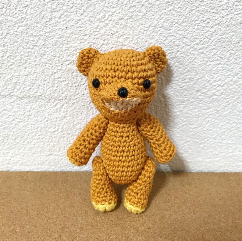 Puede incluir: Un oso de peluche de crochet con cuerpo marr&oacute;n, ojos negros y una sonrisa cosida blanca. El oso est&aacute; de pie sobre una superficie marr&oacute;n.