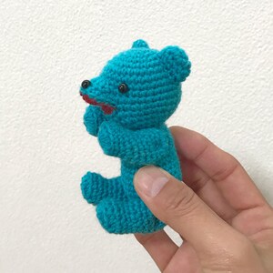 Puede incluir: Un peque&ntilde;o oso de peluche turquesa tejido a crochet con ojos negros y boca roja. El oso est&aacute; siendo sostenido en una mano.