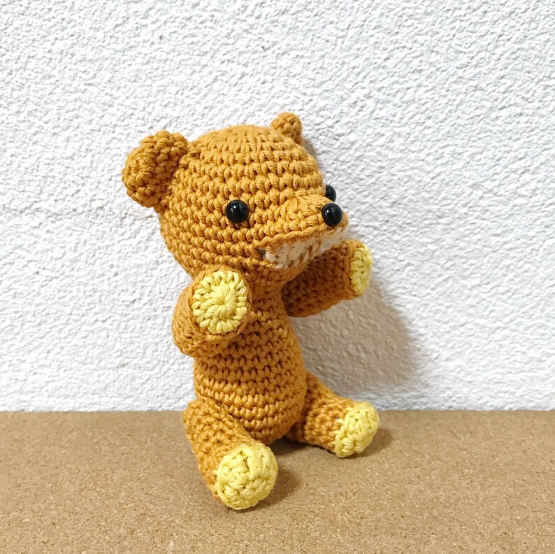 Puede incluir: Un oso de peluche de crochet con detalles amarillos y ojos negros. El oso est&aacute; sentado sobre una superficie marr&oacute;n.