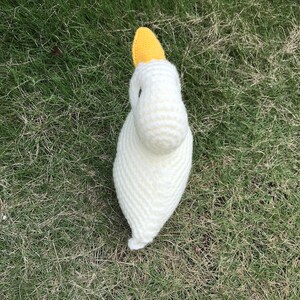 May include: A white crocheted bird with a yellow beak. The bird is sitting on a bed of green grass.