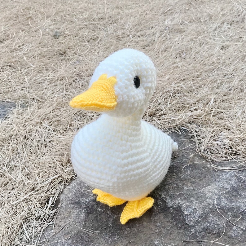 May include: A white crocheted duck with a yellow beak and feet. The duck is sitting on a brown surface.