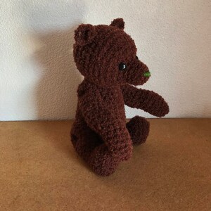 May include: A brown crocheted teddy bear with a green nose sitting on a brown surface.