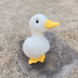 May include: A white crocheted goose with yellow feet and beak. The goose is sitting on a brown surface.