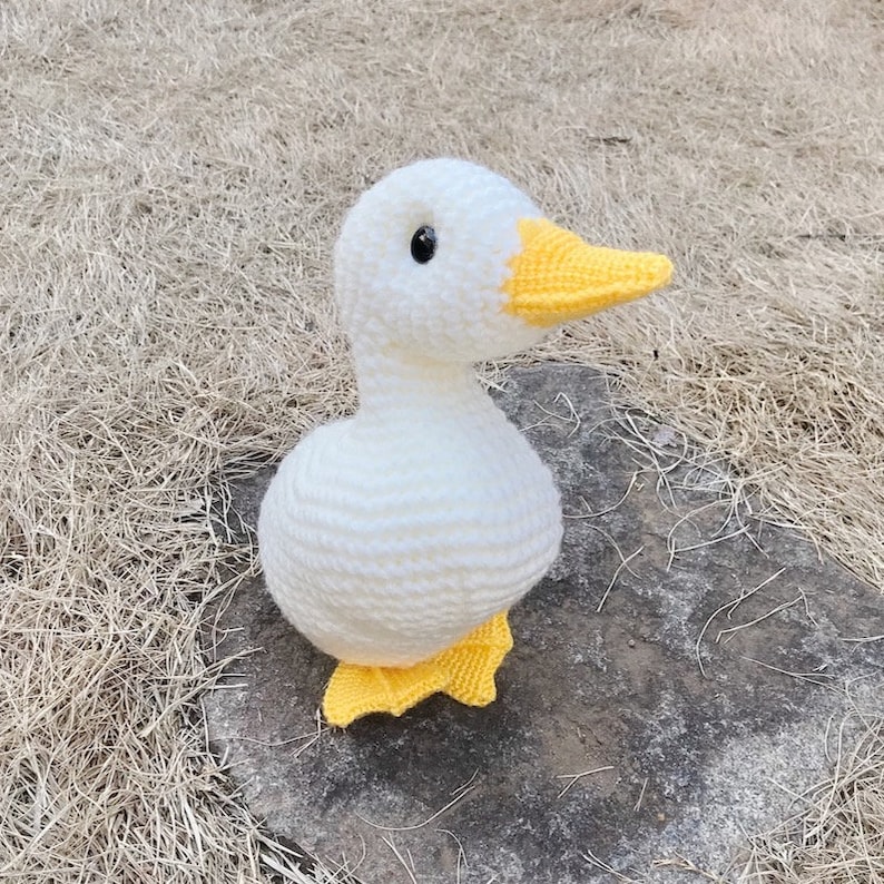 May include: A white crocheted goose with yellow feet and beak. The goose is sitting on a brown surface.