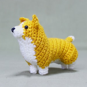 May include: A crocheted stuffed toy dog in the shape of a corgi. The dog is yellow with white accents and black eyes.