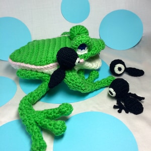 May include: A green crocheted frog with pink accents and black eyes. The frog is holding a black crocheted tadpole. There are two other black crocheted tadpoles on a blue background.