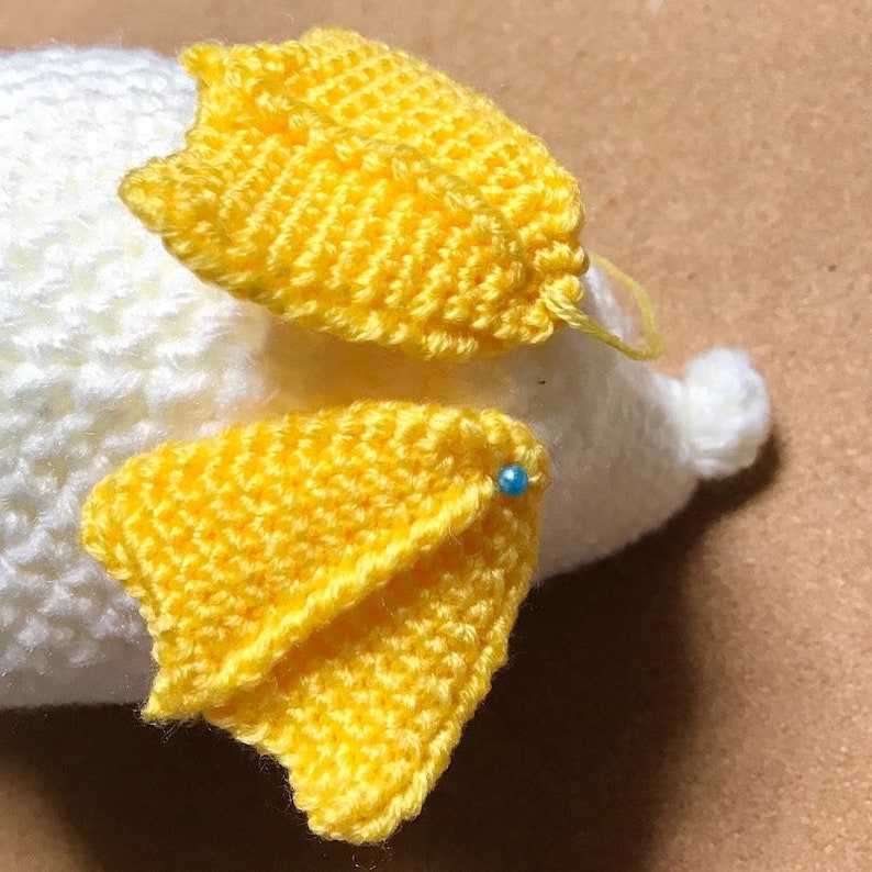 May include: Two yellow crocheted wings with a blue pin holding them in place on a white crocheted stuffed animal.