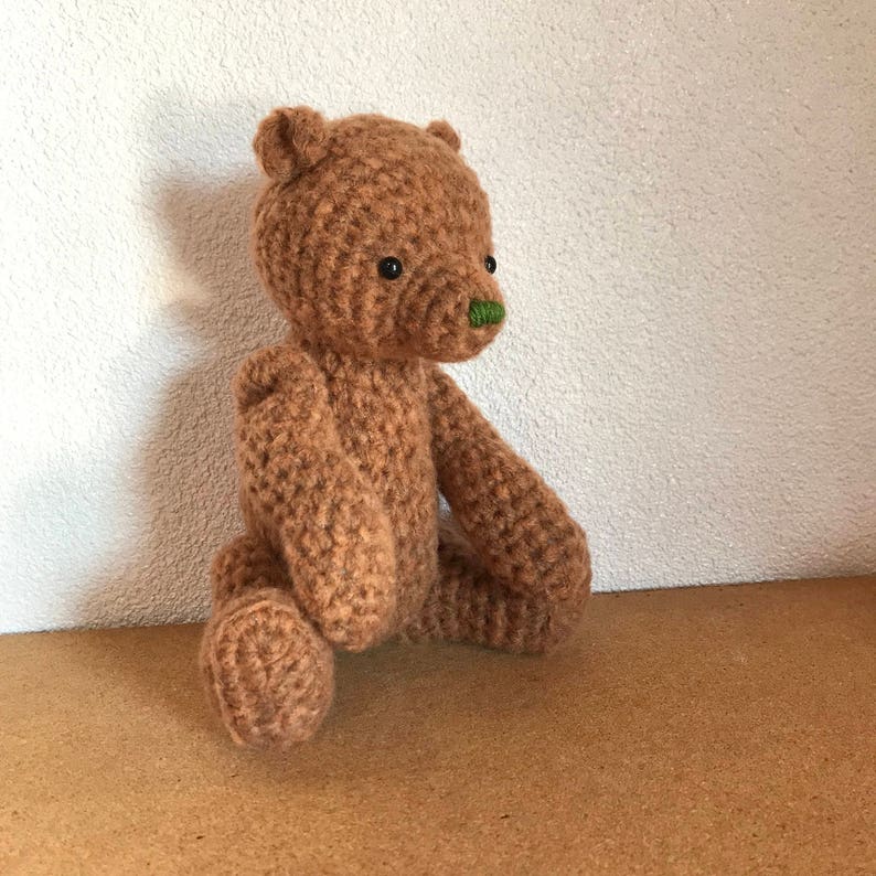 May include: A brown crocheted teddy bear with green embroidered nose sitting on a brown surface.