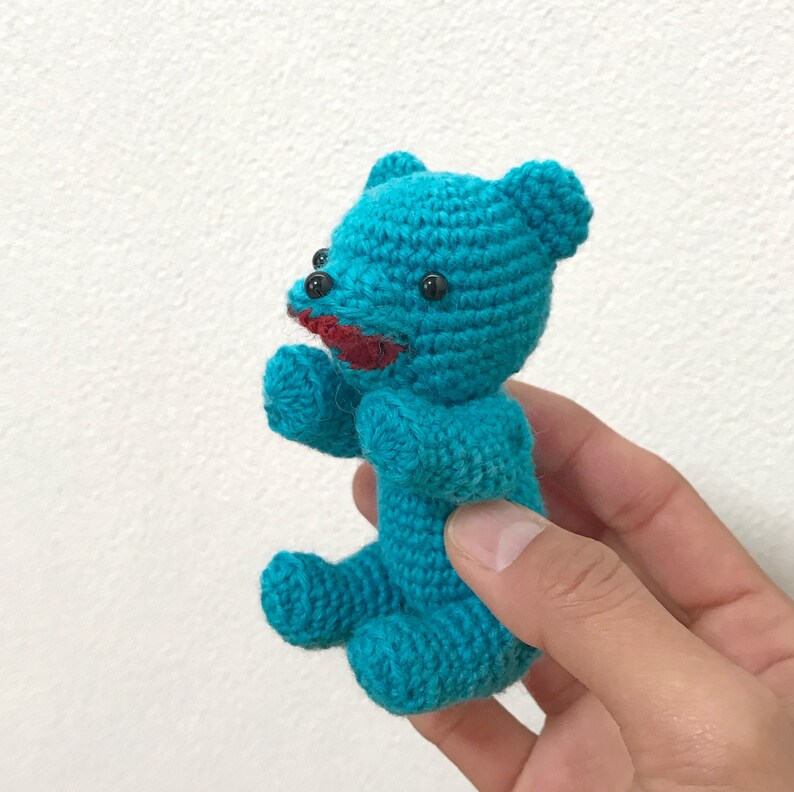 Puede incluir: Un peque&ntilde;o osito de peluche de crochet azul turquesa con ojos negros y boca roja. El oso est&aacute; siendo sostenido en la mano de una persona.