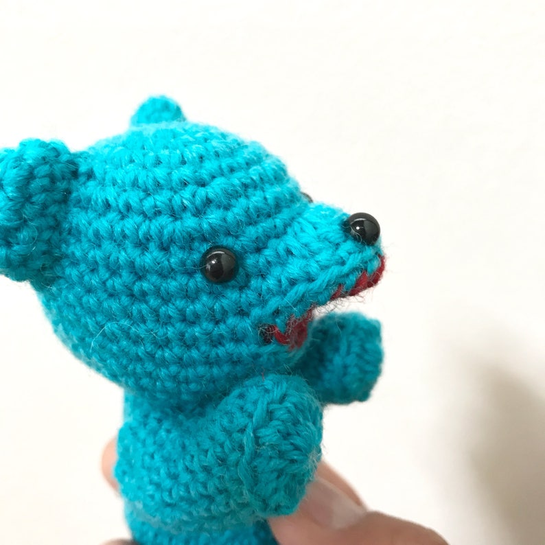 Puede incluir: Un oso de peluche de crochet azul turquesa con ojos negros y boca roja. El oso mira hacia la derecha.