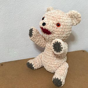 Puede incluir: Un oso de peluche de crochet con pelaje rosa y blanco, labios rojos y ojos negros y grises. El oso est&aacute; sentado sobre una superficie marr&oacute;n.