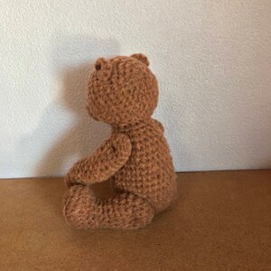 May include: A brown crocheted teddy bear sitting on a brown surface. The bear is facing away from the camera and has its arms crossed.