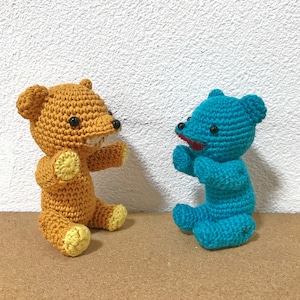 Puede incluir: Dos osos de peluche tejidos a crochet, uno marr&oacute;n con detalles amarillos y otro azul. El oso marr&oacute;n tiene una sonrisa de dientes y el oso azul tiene una boca roja.
