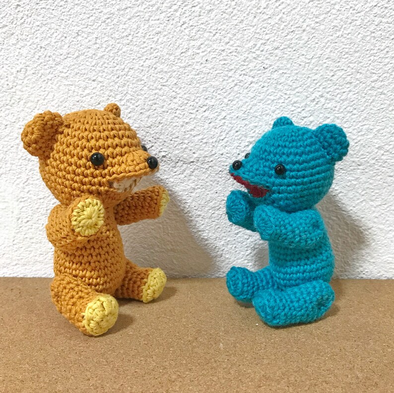 Puede incluir: Dos osos de peluche tejidos a crochet, uno marr&oacute;n con detalles amarillos y otro azul. El oso marr&oacute;n tiene una sonrisa de dientes y el oso azul tiene una boca roja.