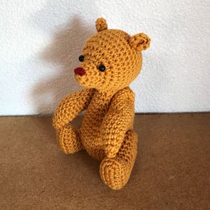 May include: A crocheted teddy bear with brown fur, black eyes, and a red nose. The bear is sitting on a brown surface.