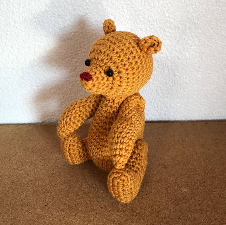 May include: A crocheted teddy bear with brown fur, black eyes, and a red nose. The bear is sitting on a brown surface.