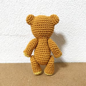 Puede incluir: Un oso de peluche de crochet con pelaje marr&oacute;n y patas amarillas. El oso est&aacute; de pie sobre una superficie marr&oacute;n.