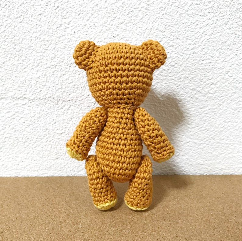 Puede incluir: Un oso de peluche de crochet con pelaje marr&oacute;n y patas amarillas. El oso est&aacute; de pie sobre una superficie marr&oacute;n.