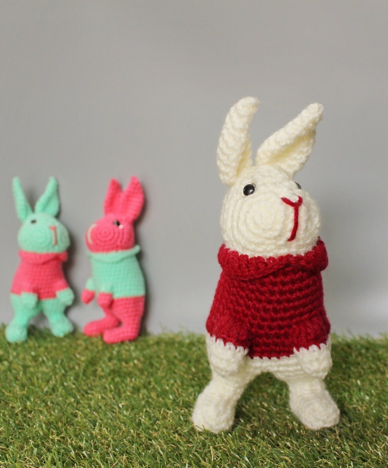May include: Three crocheted bunny rabbits. One is white with a red sweater, one is pink with a green sweater, and one is green with a pink sweater.