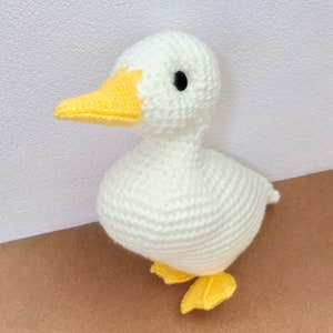 May include: A white crocheted duck with yellow beak and feet. The duck is standing on a brown surface.