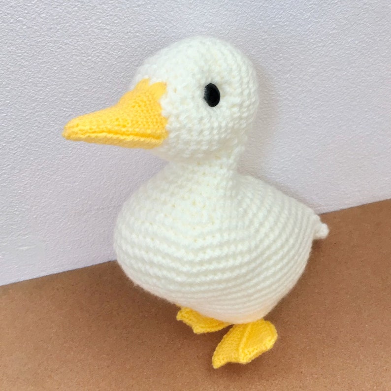 May include: A white crocheted duck with yellow beak and feet. The duck is standing on a brown surface.