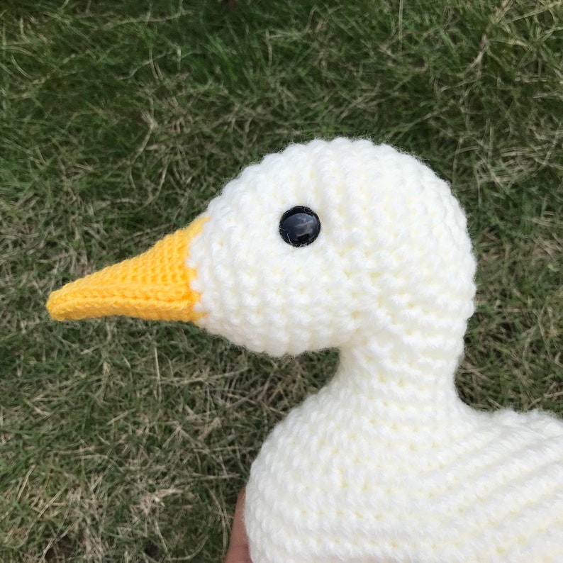 May include: Close-up of a white crocheted goose with a yellow beak and black eyes. The goose is sitting on a green grassy surface.